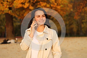 beautiful caucasian girl with dark hair in a raincoat stands on a background of autumn trees holding a smartphone in her hand