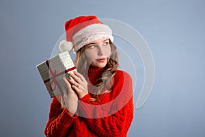 Beautiful Caucasian girl in dress and Santa hat is holding presents, looking at camera and smiling, isolated on gray background