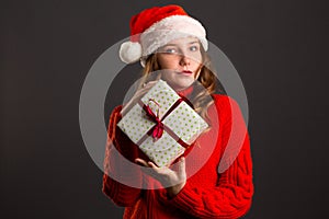 Beautiful Caucasian girl in dress and Santa hat is holding presents, looking at camera and smiling, isolated on gray background