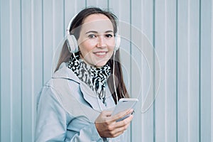 Beautiful caucasian girl in a gray coat listens to music on headphones through a smartphone