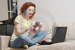 Beautiful caucasian girl sitting on the sofa at home, listening to music on headphones and using a smartphone.