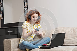 Beautiful caucasian girl sitting on the sofa at home, listening to music on headphones and using a smartphone.