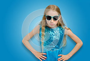 Beautiful Caucasian little girl in sunglasses and blue dress looks angrily into the camera. Emotions and adolescent photo