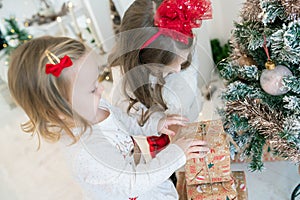 Beautiful Caucasian little girls wearing festive outfits while opening gift boxes from Santa Claus found under the Christmas tree