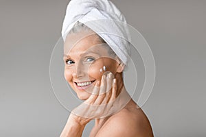 Beautiful caucasian mature woman applying moisturizing cream on face posing with towel on head and smiling at camera