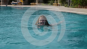 Beautiful Caucasian model, young woman is swimming in a pool with blue water in a hotel, under the open sky. The concept