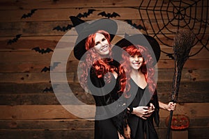 Beautiful caucasian mother and her daughter with long red hair in witch costumes celebrating Halloween posing with over
