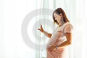 Beautiful caucasian pregnant woman touching her big belly in front of the window behind the curtain. Expectation of the childbirth