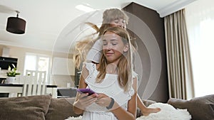 Beautiful caucasian student girl sits on bed using smartphone and smiling her little sister have fun jumps behind her