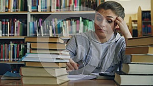 Beautiful caucasian teenage woman is sitting at table in university library holding digital tablet. She is tired and