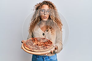 Beautiful caucasian teenager girl eating tasty pepperoni pizza clueless and confused expression