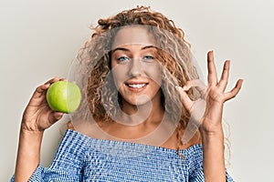 Beautiful caucasian teenager girl holding green apple doing ok sign with fingers, smiling friendly gesturing excellent symbol