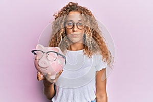 Beautiful caucasian teenager girl holding piggy bank with glasses thinking attitude and sober expression looking self confident