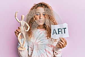 Beautiful caucasian teenager girl holding wooden manikin and art word relaxed with serious expression on face