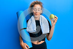 Beautiful caucasian teenager girl holding yoga mat and green apple looking at the camera blowing a kiss being lovely and sexy