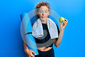 Beautiful caucasian teenager girl holding yoga mat and green apple relaxed with serious expression on face