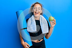 Beautiful caucasian teenager girl holding yoga mat and green apple smiling and laughing hard out loud because funny crazy joke