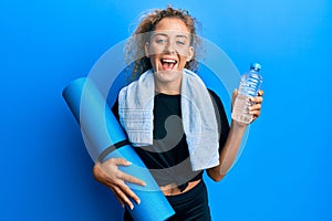 Beautiful caucasian teenager girl holding yoga mat and water bottle smiling and laughing hard out loud because funny crazy joke