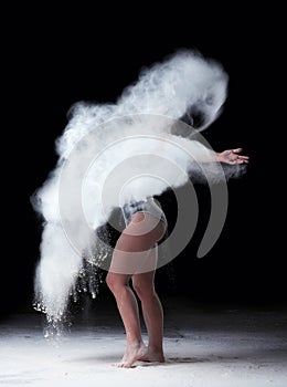 Beautiful caucasian woman in a black bodysuit with a sports figure is dancing in a white cloud of flour