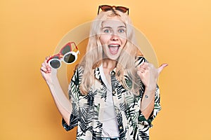 Beautiful caucasian woman with blond hair holding many sunglasses pointing thumb up to the side smiling happy with open mouth