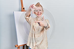 Beautiful caucasian woman with blond hair standing by painter easel stand smiling making frame with hands and fingers with happy
