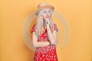 Beautiful caucasian woman with blond hair wearing summer hat with hand on chin thinking about question, pensive expression