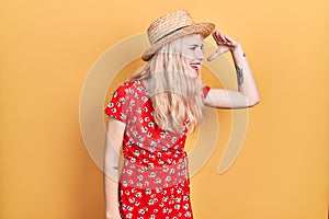 Beautiful caucasian woman with blond hair wearing summer hat very happy and smiling looking far away with hand over head