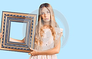 Beautiful caucasian woman with blonde hair holding empty frame thinking attitude and sober expression looking self confident