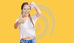 Beautiful caucasian woman with blonde hair wearing braids and white tshirt smiling making frame with hands and fingers with happy