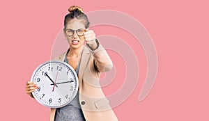 Beautiful caucasian woman with blonde hair wearing business jacket and holding clock annoyed and frustrated shouting with anger,