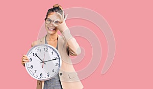 Beautiful caucasian woman with blonde hair wearing business jacket and holding clock smiling happy doing ok sign with hand on eye