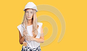 Beautiful caucasian woman with blonde hair wearing hardhat and builder clothes thinking attitude and sober expression looking self
