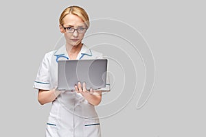 Beautiful Caucasian woman doctor or nurse holding a laptop pc computer standing over grey background