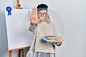 Beautiful caucasian woman drawing with palette on easel stand winner first place with open hand doing stop sign with serious and