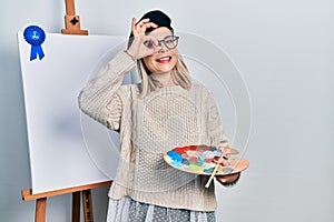 Beautiful caucasian woman drawing with palette on easel stand winner first place smiling happy doing ok sign with hand on eye photo