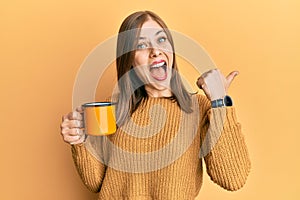 Beautiful caucasian woman drinking a cup coffee pointing thumb up to the side smiling happy with open mouth