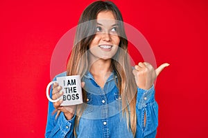 Beautiful caucasian woman drinking from i am the boss coffee cup pointing thumb up to the side smiling happy with open mouth