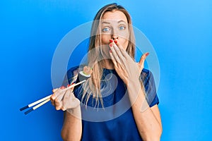 Beautiful caucasian woman eating temaki sushi using chopsticks covering mouth with hand, shocked and afraid for mistake