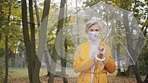 Beautiful caucasian woman with face mask holding with transparent umbrela in the park in autumn