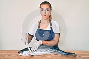 Beautiful caucasian woman fishmonger selling fresh raw salmon in shock face, looking skeptical and sarcastic, surprised with open