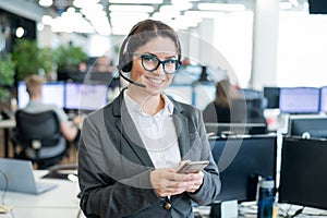 Beautiful caucasian woman in headset is holding a mobile phone while standing in open space office. Friendly female