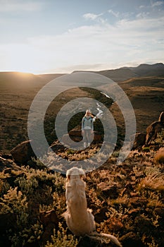 Beautiful Caucasian woman and her dog on top of the mountain enjoying the view during sunset