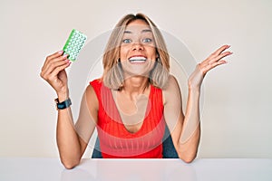 Beautiful caucasian woman holding birth control pills celebrating victory with happy smile and winner expression with raised hands