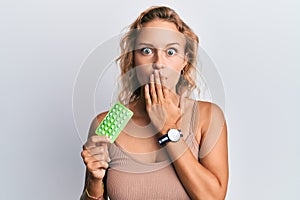 Beautiful caucasian woman holding birth control pills covering mouth with hand, shocked and afraid for mistake