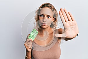 Beautiful caucasian woman holding birth control pills with open hand doing stop sign with serious and confident expression,