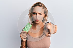 Beautiful caucasian woman holding birth control pills pointing with finger to the camera and to you, confident gesture looking