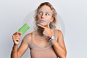 Beautiful caucasian woman holding birth control pills serious face thinking about question with hand on chin, thoughtful about
