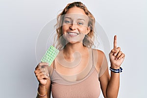 Beautiful caucasian woman holding birth control pills surprised with an idea or question pointing finger with happy face, number