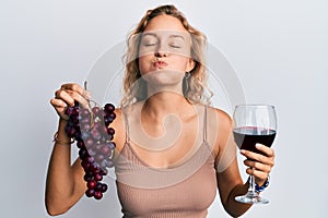 Beautiful caucasian woman holding branch of fresh grapes and red wine puffing cheeks with funny face