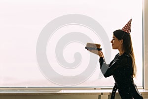 beautiful caucasian woman holding a cake looking at a candle, standing at the office window, light background, profile